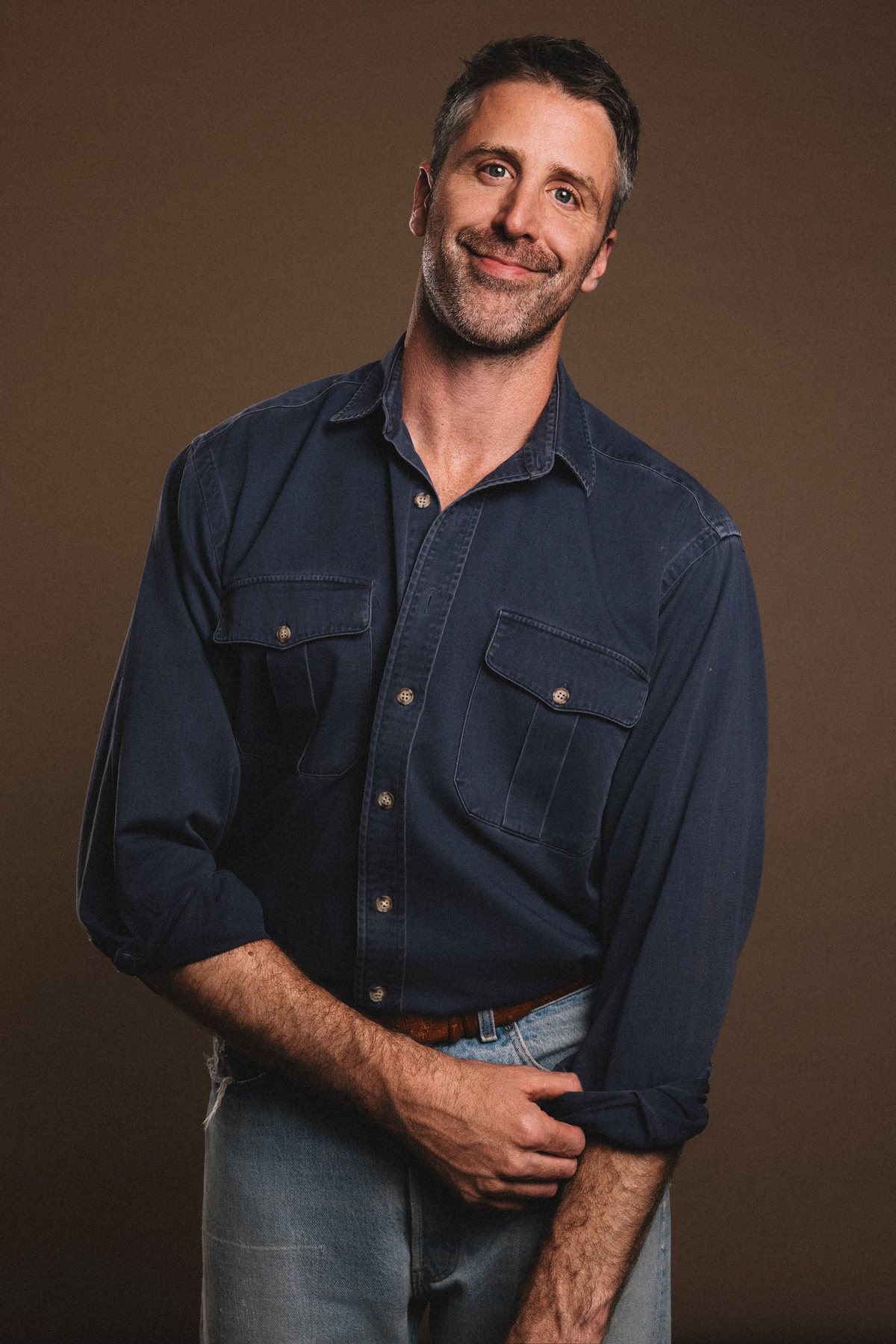 Jason S. Burke in a navy work shirt and jeans, smiling, against a brown backdrop